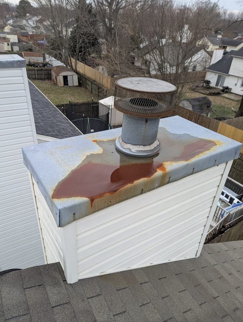 Rusted metal chimney chase top with a round flue cap on a white-sided chimney