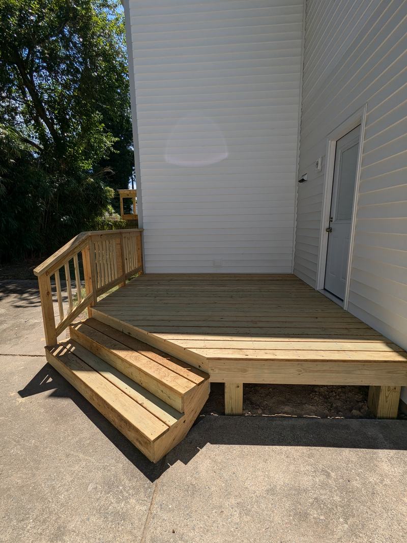 Ground-level wood deck with wide steps and a wood side railing against white vinyl siding beside a side door