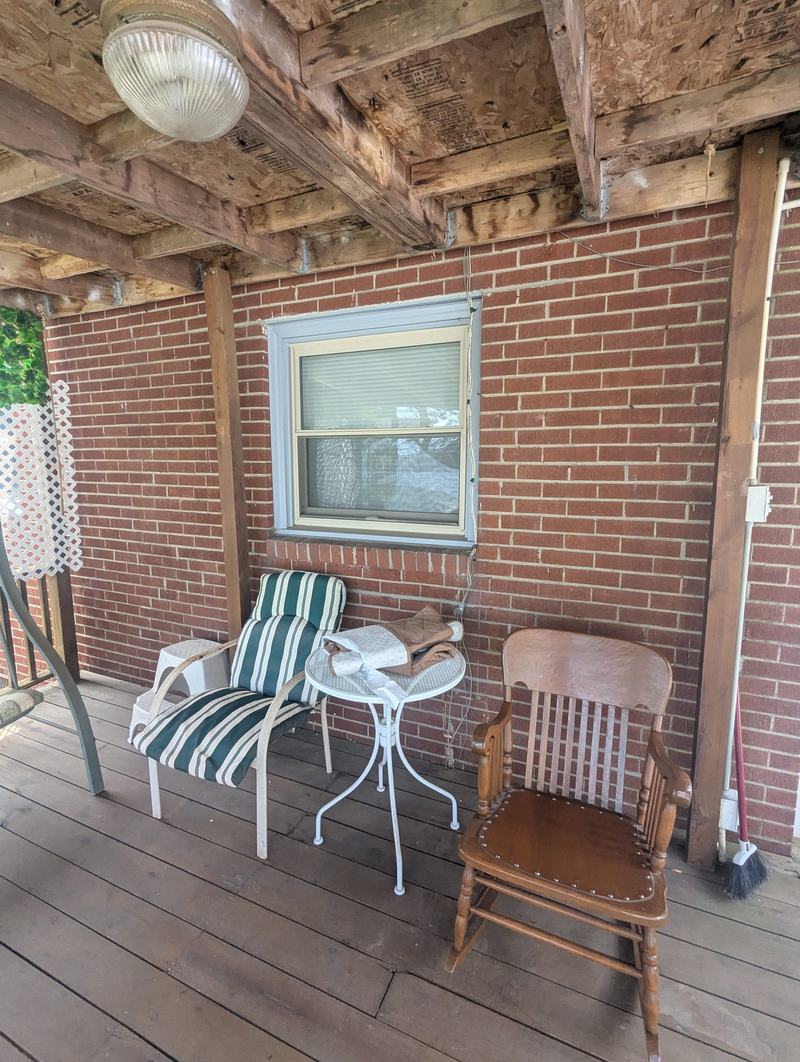 Brick wall with a single window on a covered wood-deck porch with patio chairs
