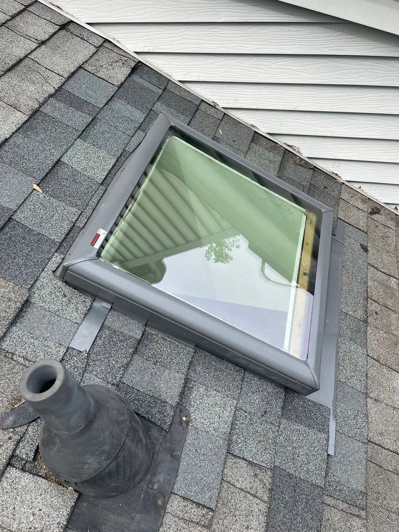 Skylight set in gray asphalt shingles with metal flashing, plumbing vent in the foreground